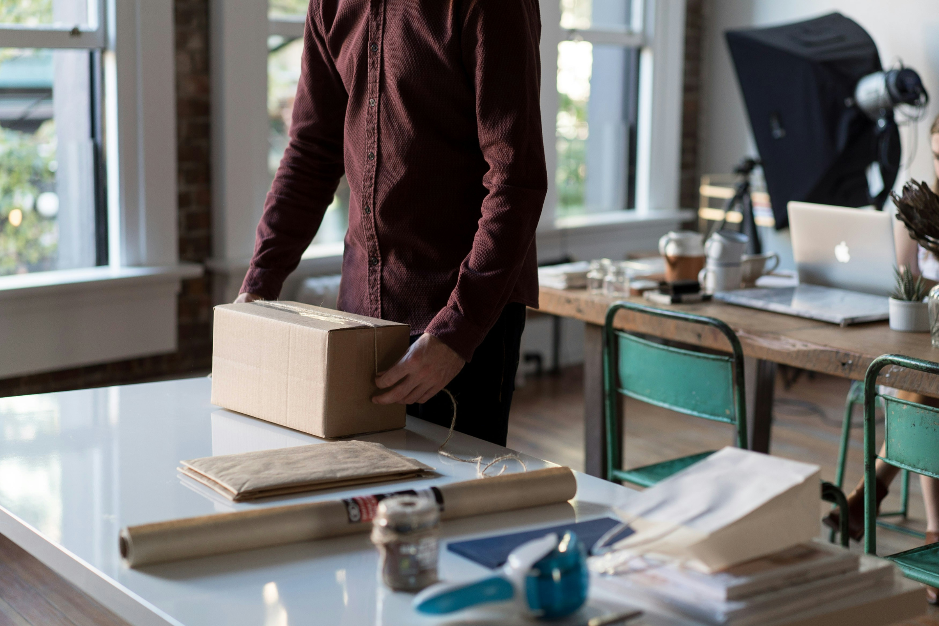 A small business owner wearing a dark red shirt stands at a table preparing a package for shipping
