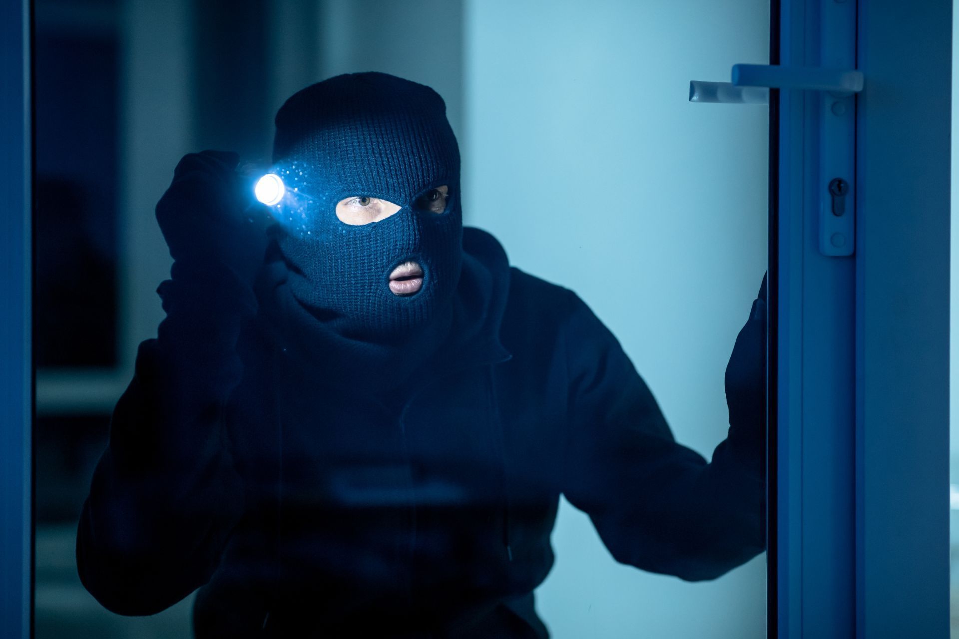 A burglar dressed in all-black clothes peers into the window at night while holding a flashlight
