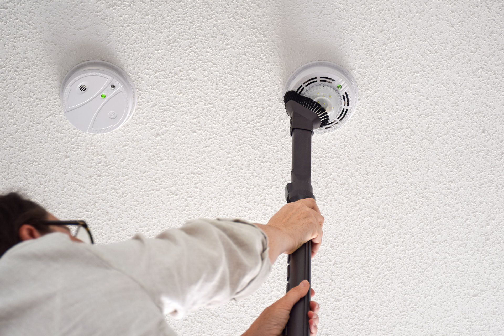 A person gently cleaning a carbon monoxide alarm using a vacuum with a brush attachment.