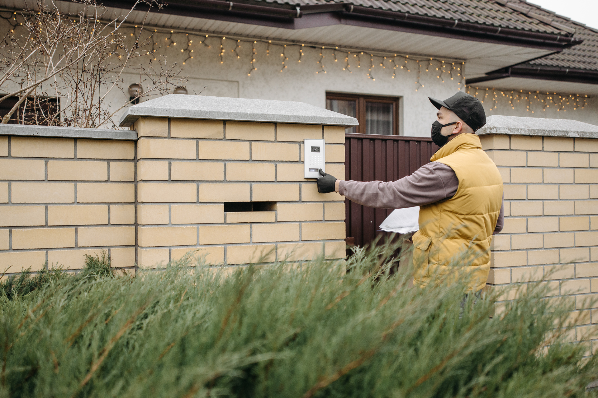 A delivery person wearing a yellow vest, a mask, and a black baseball cap ringing a video doorbell at the front gate of a house