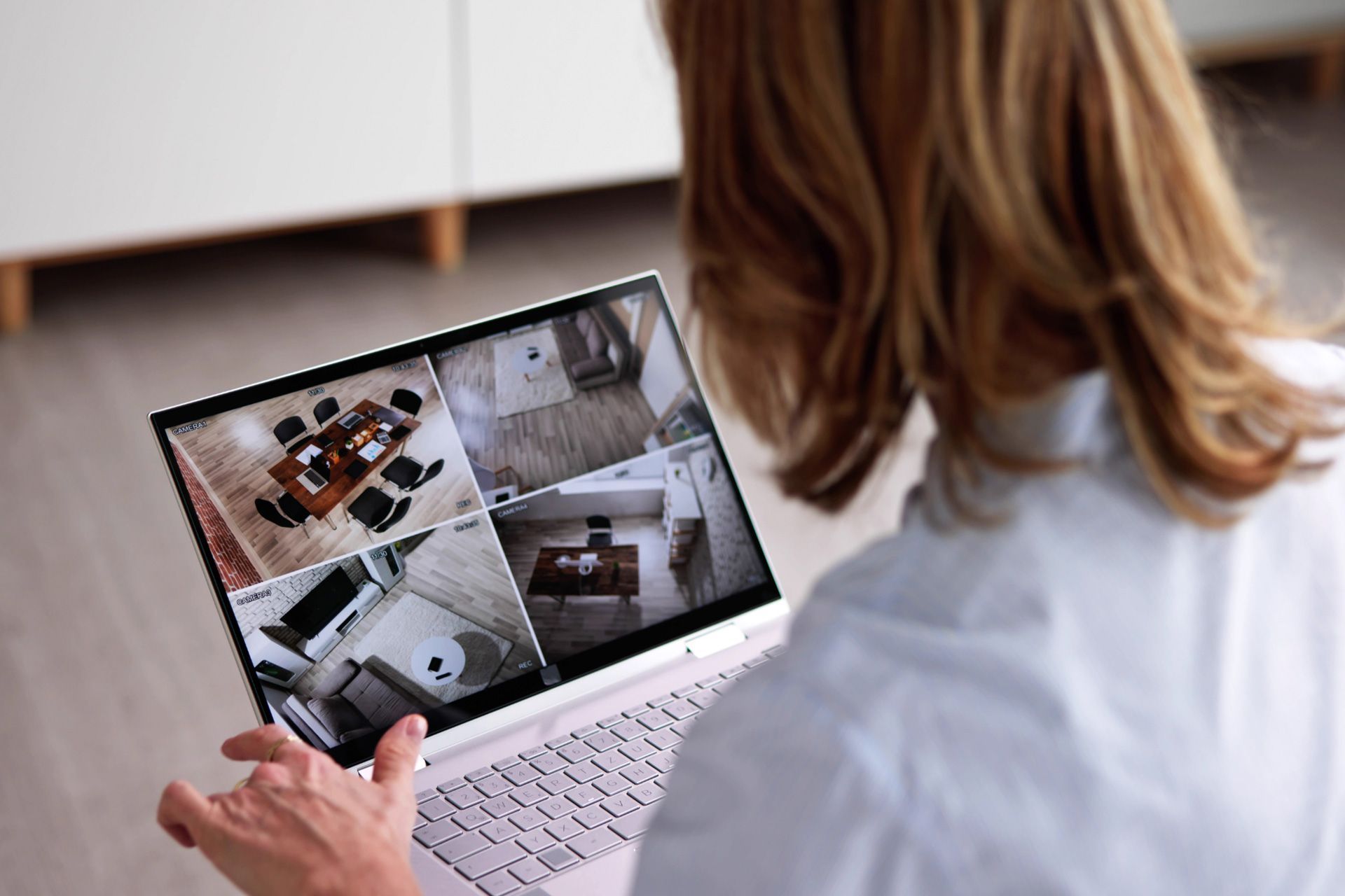 A woman monitors her home from a laptop with footage from indoor security cameras
