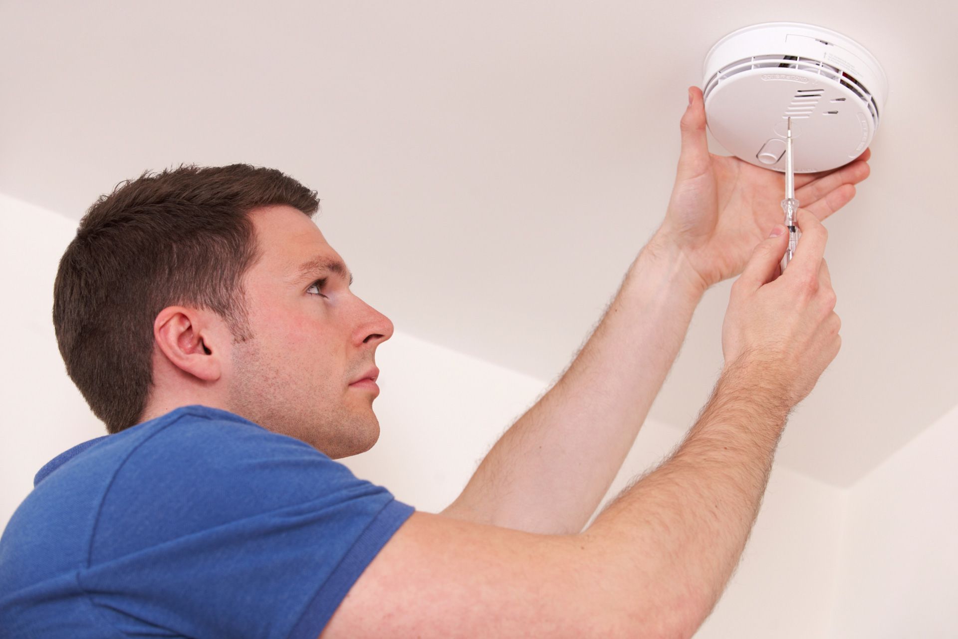 A man wearing a blue T-shirt replaces a smoke alarm on the ceiling