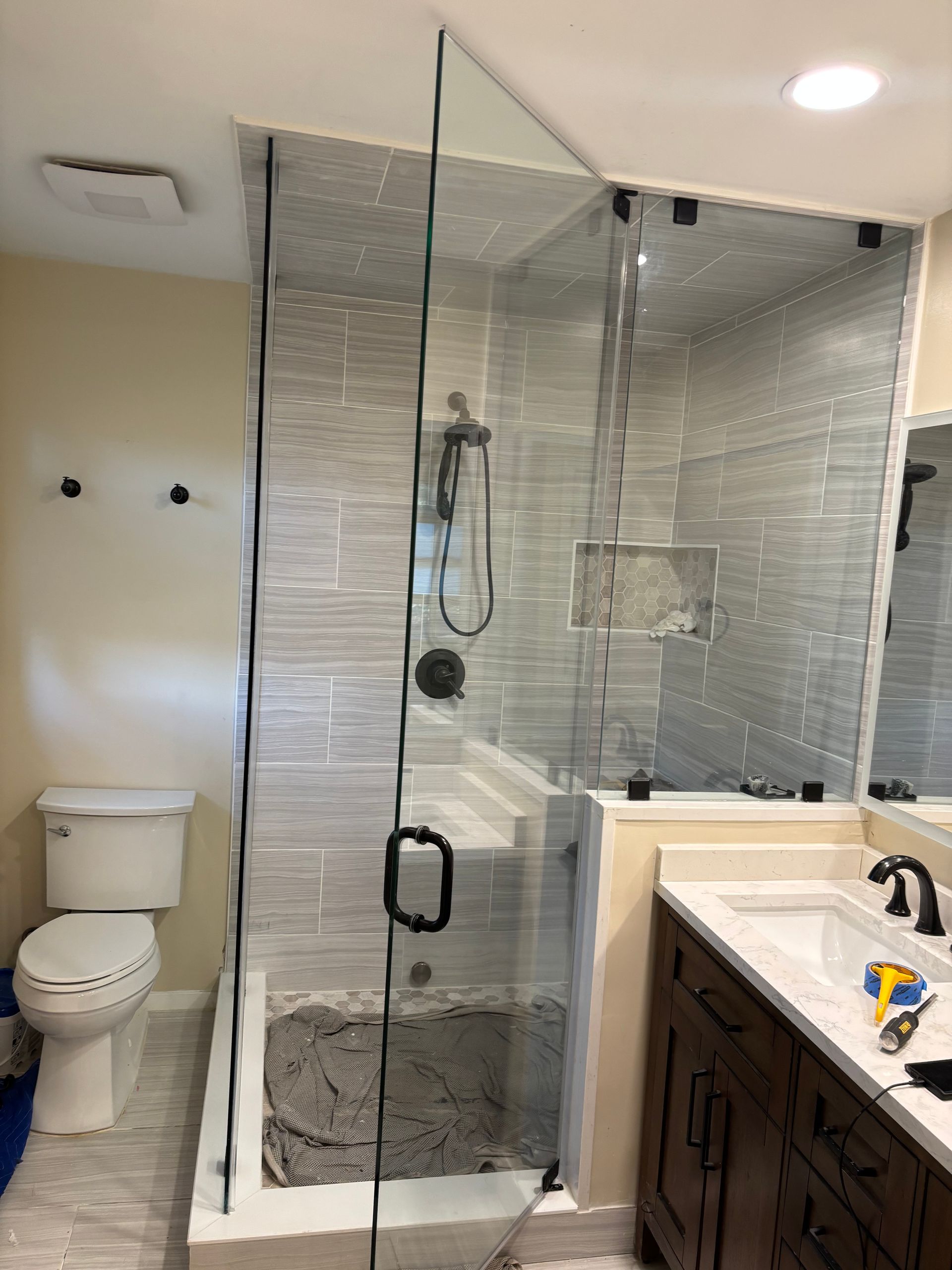 Bathroom with glass shower enclosure, gray tiled walls, toilet, vanity with sink, and dark brown cabinet.