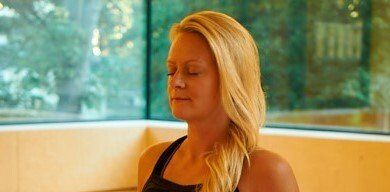 Woman with closed eyes meditating, blonde hair, black top, bright room with window.