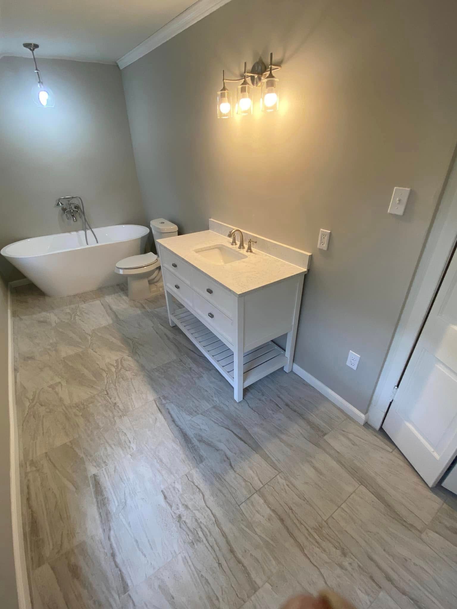 A modern bathroom with a freestanding white tub, a white vanity cabinet, a wall-mounted light fixture, and tiled flooring.