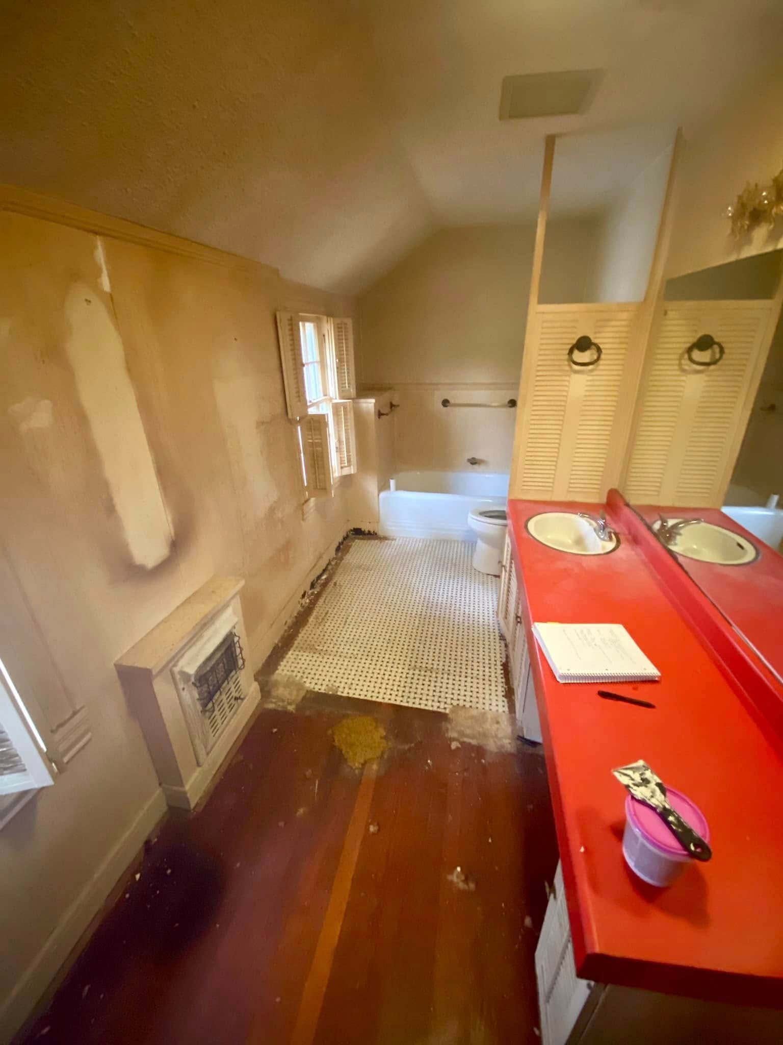 A bathroom undergoing renovation with patched beige walls, a red vanity countertop, and a white patterned floor rug.