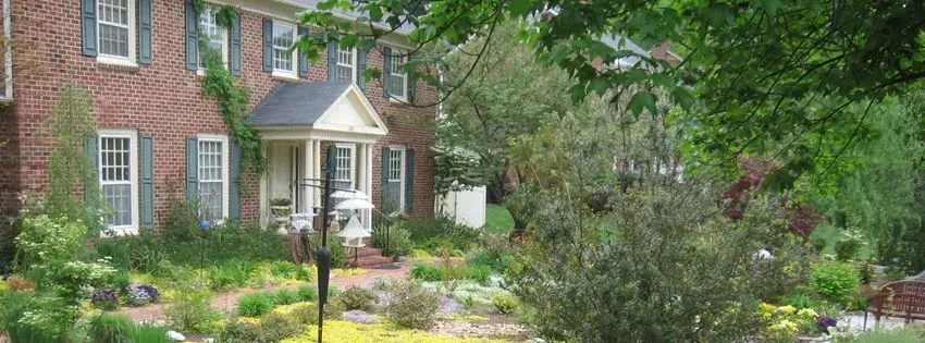 Brick house with white trim, green shutters, and a well-manicured yard with trees and bushes.
