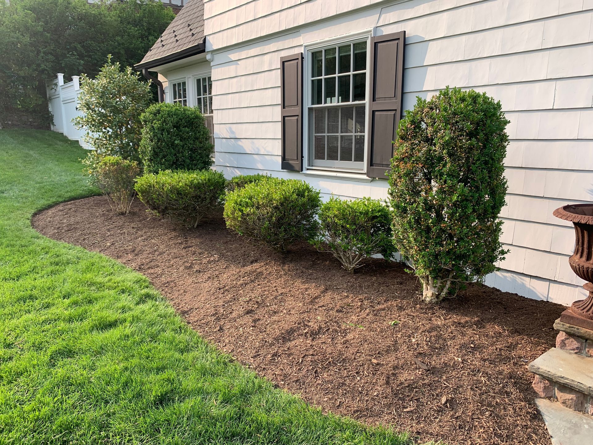 Brown mulch bed with green bushes and lawn along a white house with brown shutters.