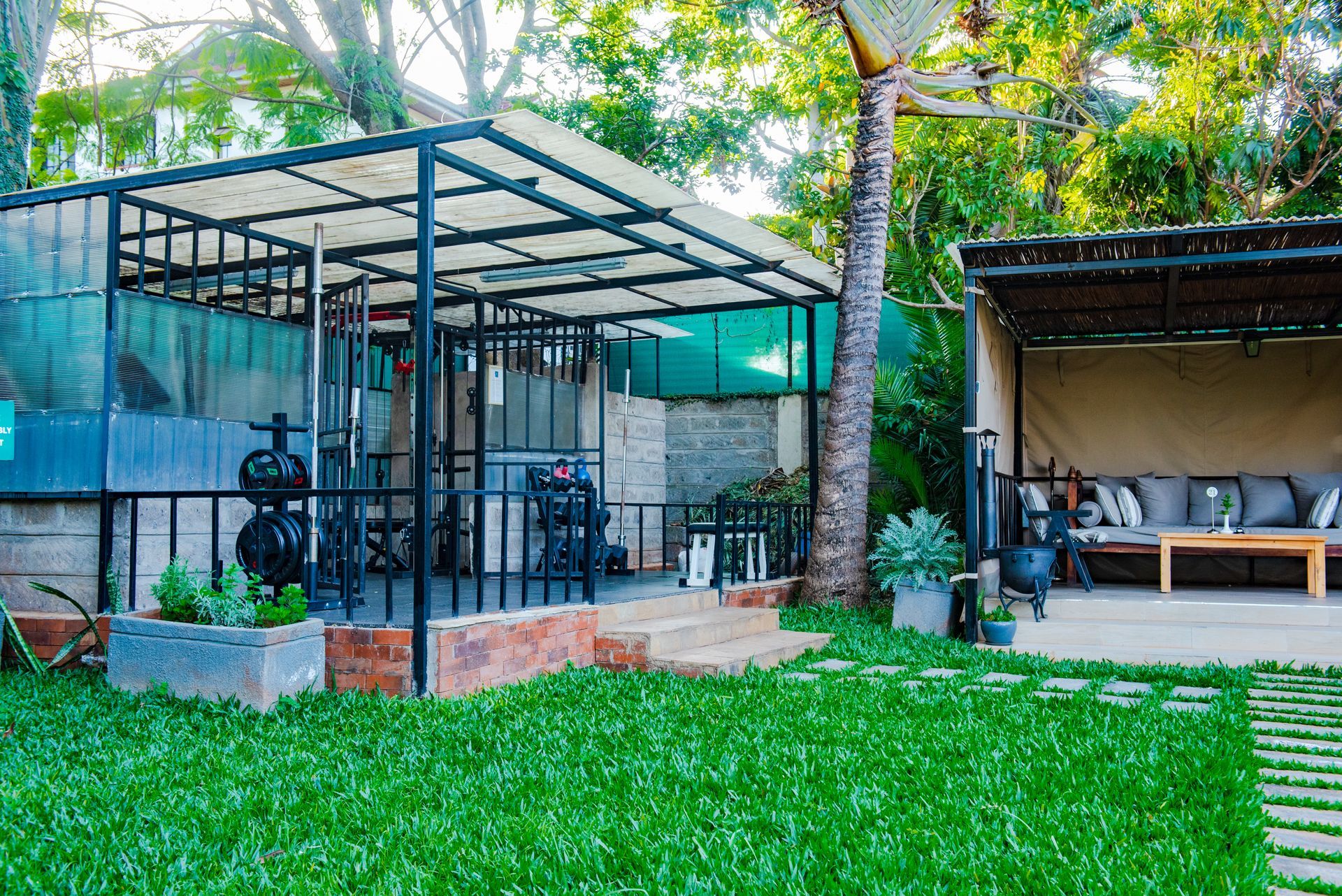 Outdoor gym with weights and equipment, and a separate covered lounge area with seating, all surrounded by greenery.