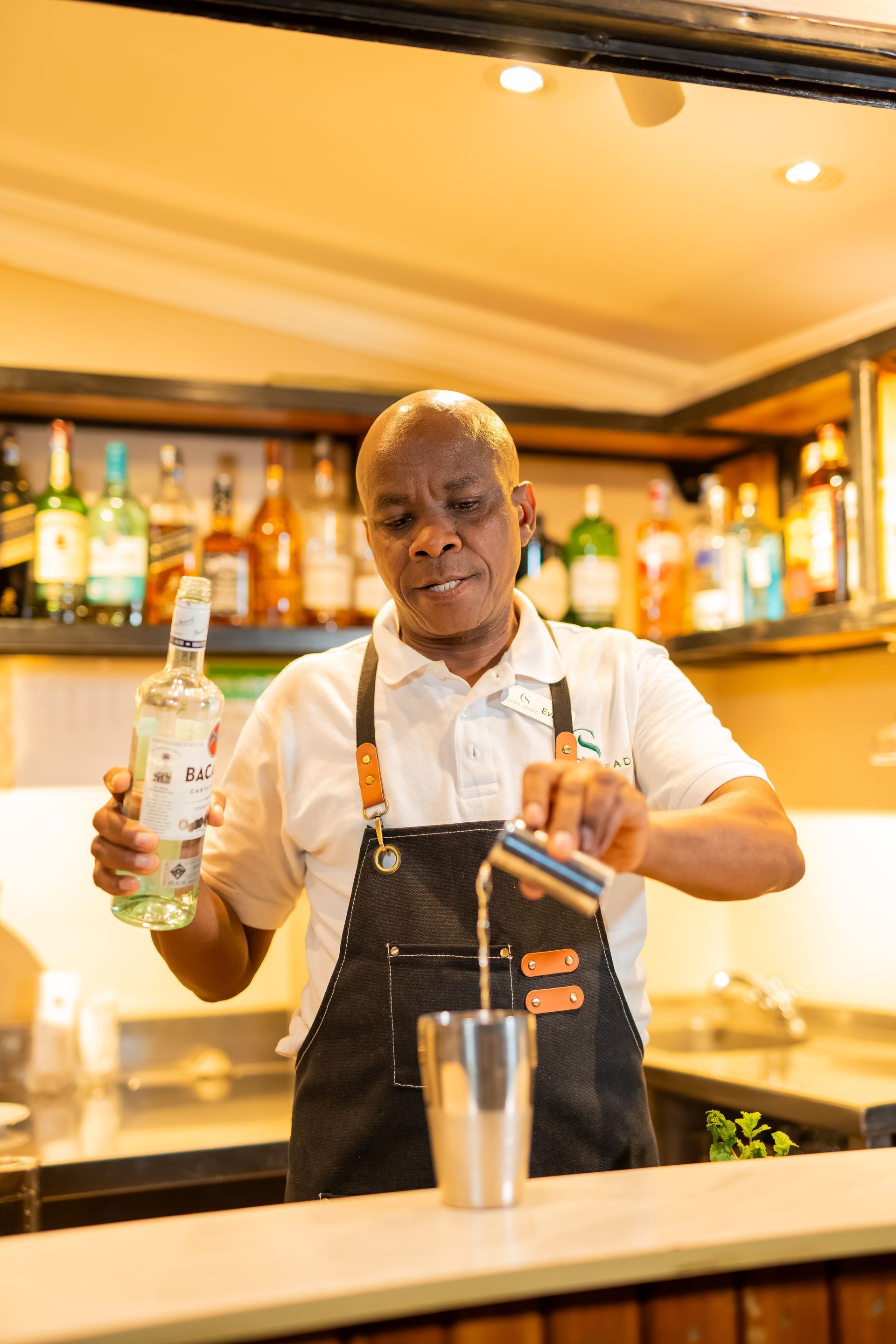 Bartender pouring a drink into a shaker. He wears a black apron and stands behind a bar with bottles on a shelf.