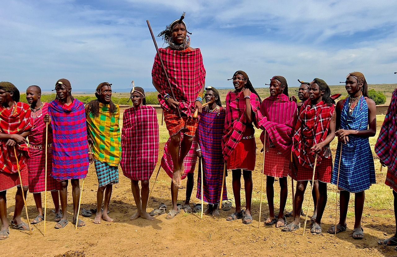 Group of Maasai men in colorful robes; one jumps high, others hold sticks. Outdoors, sunny day.