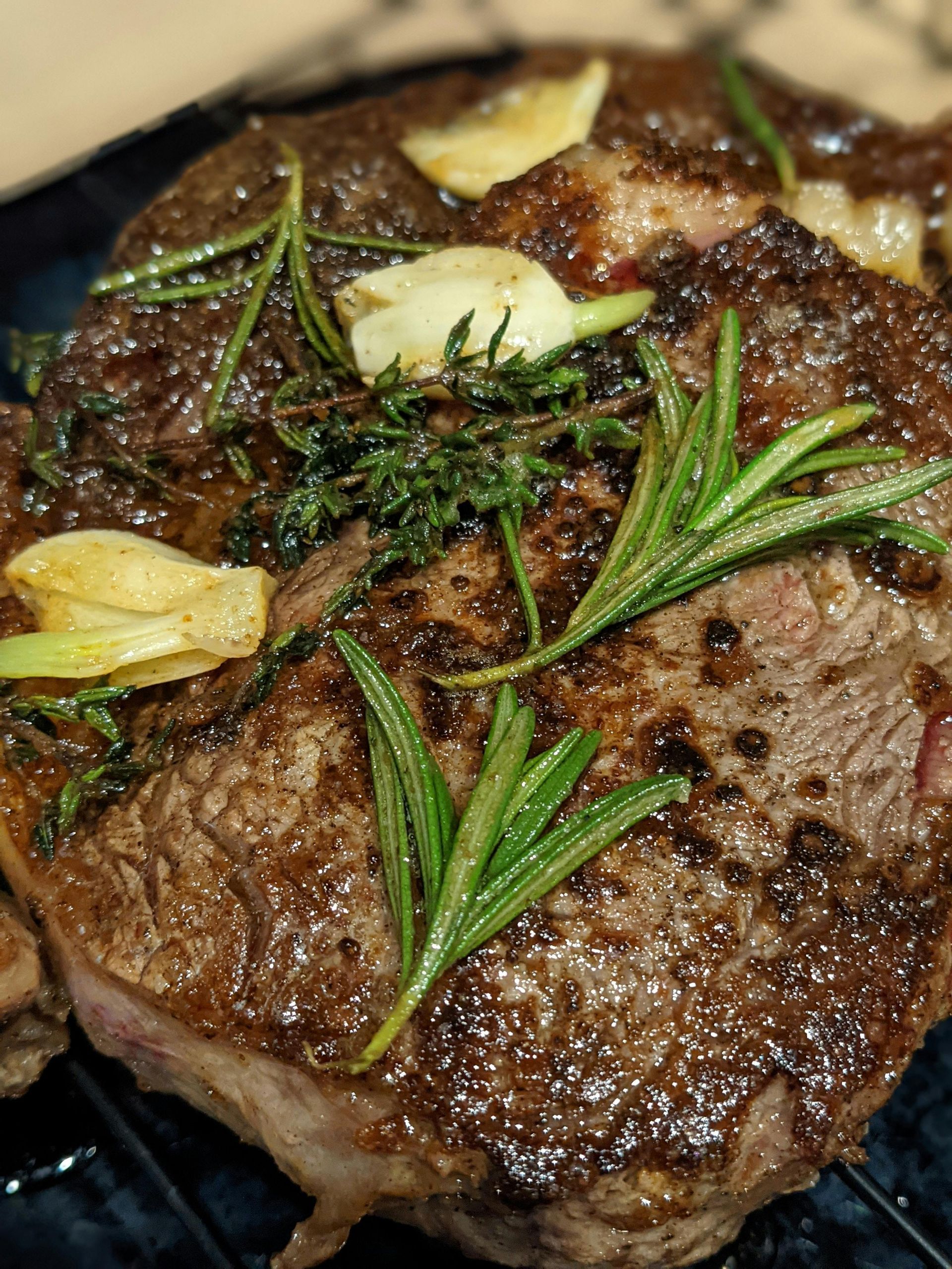Close-up of a cooked ribeye steak, seasoned with rosemary sprigs, garlic, and thyme. The steak is on a dark serving dish.