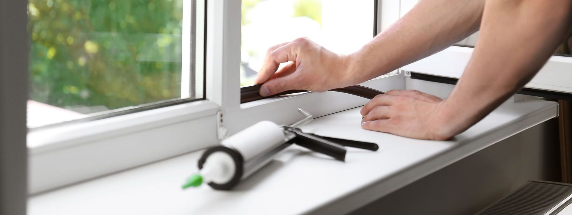 Une personne calfeutre un cadre de fenêtre ; le pistolet à calfeutrer est sur un rebord de fenêtre.