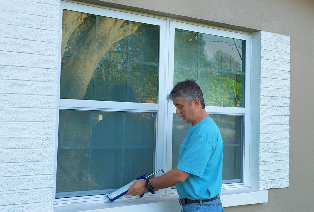 Un homme en chemise bleue applique du mastic sur une fenêtre.