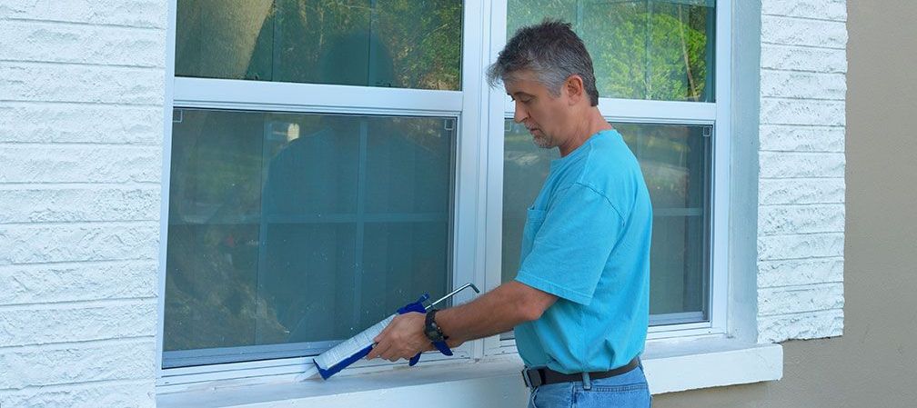 Un homme en chemise bleue applique du mastic sur une fenêtre.