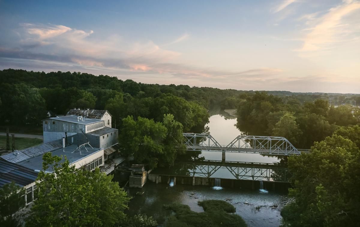 Building and bridge over a river, trees surround, sunset colors.