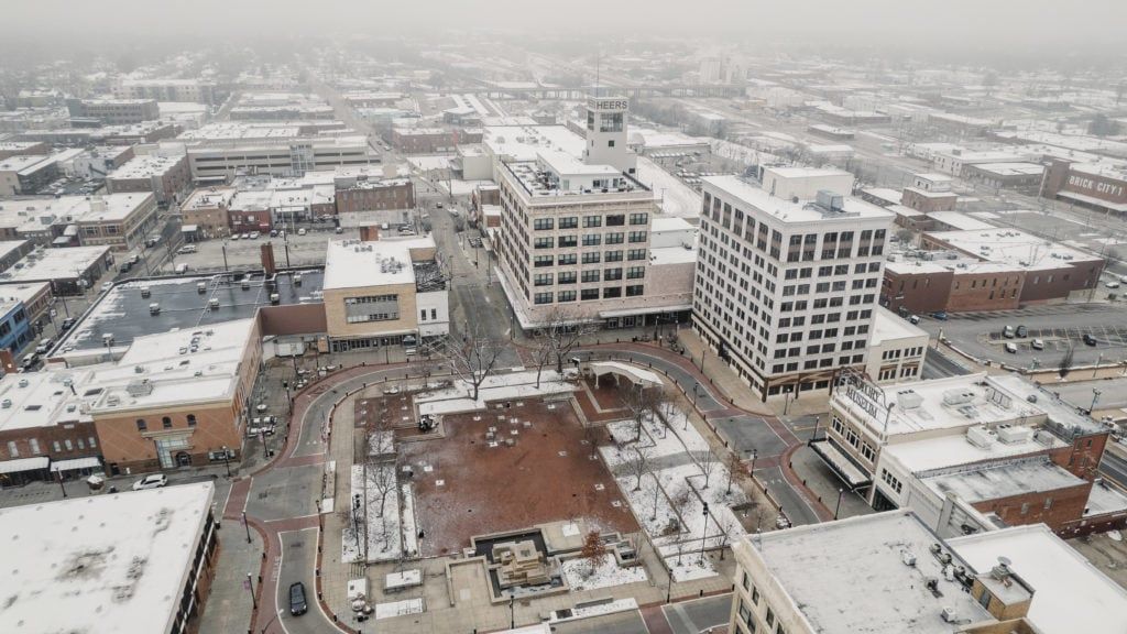 Snowy aerial view of a town square, buildings, and streets covered in snow.