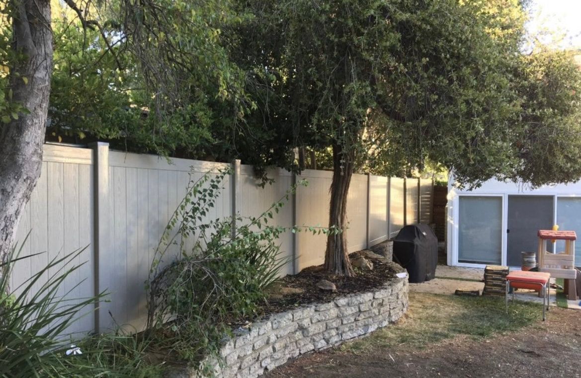 A white fence surrounds a backyard with trees and a stone wall.
