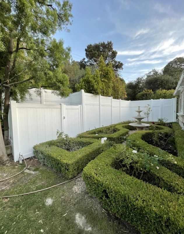 A white fence surrounds a garden with bushes and a fountain.