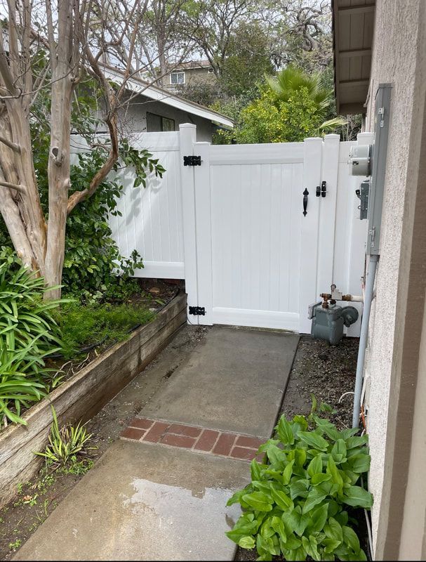 A white fence with a black handle is surrounded by plants and trees.