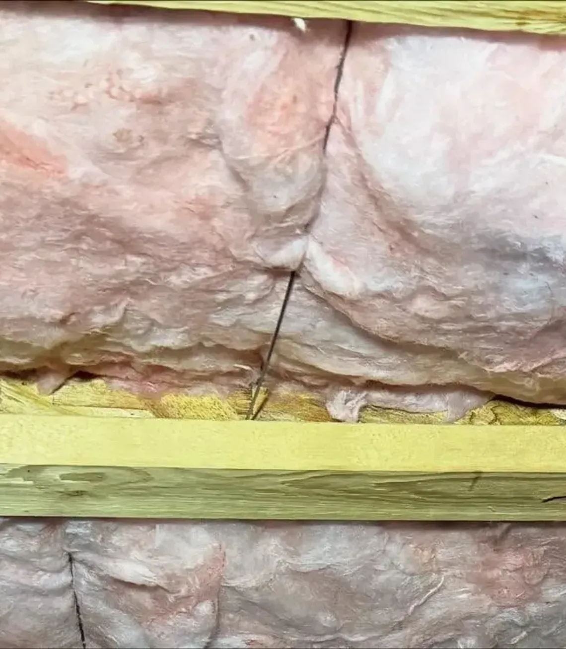 Pink fiberglass insulation installed between wooden ceiling joists, secured by thin metal wire supports.