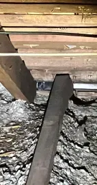 A view inside an attic showing wooden support beams and dusty, grey insulation between rafters.
