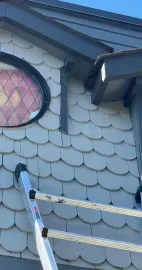 A ladder leans against a wall covered in light-colored scalloped shingles, beneath a round stained-glass window.