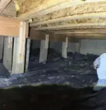 A person in a white hazmat suit and respirator examines a dark, muddy crawlspace with wooden floor joists above.