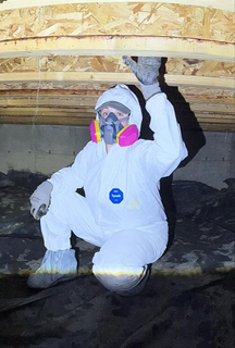 A person in a white hazmat suit and respirator examines a dark, muddy crawlspace with wooden floor joists above.