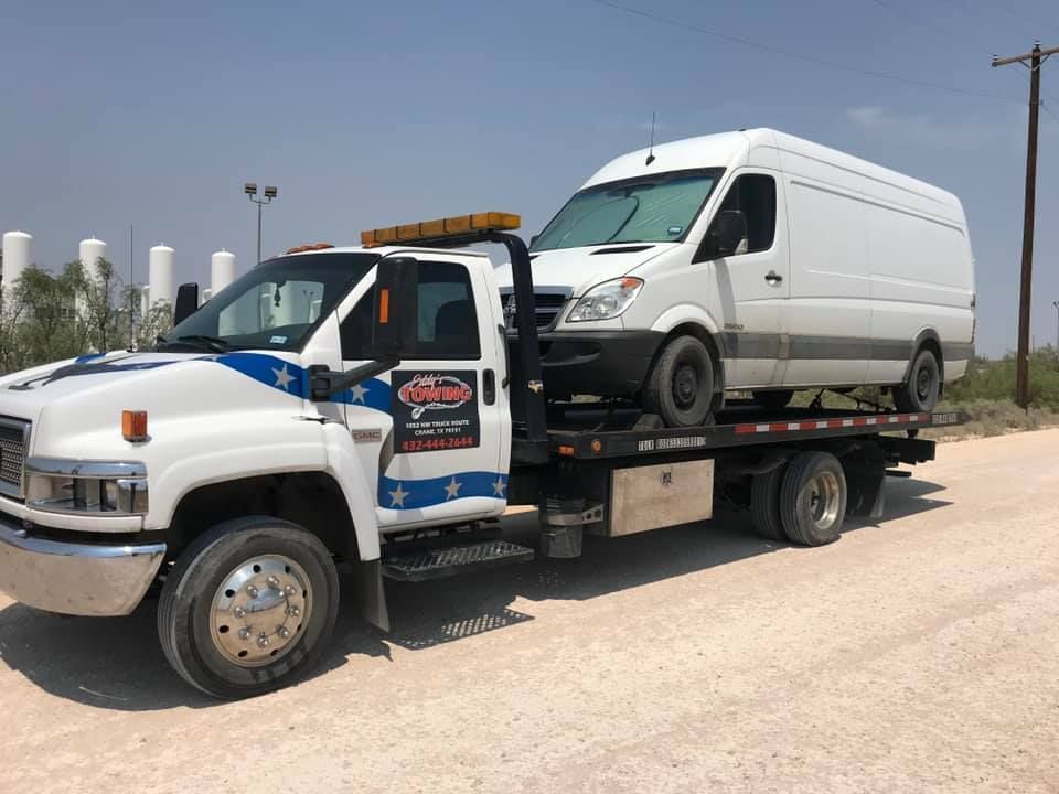 Tow truck carrying a white van on a dirt road under a sunny sky.