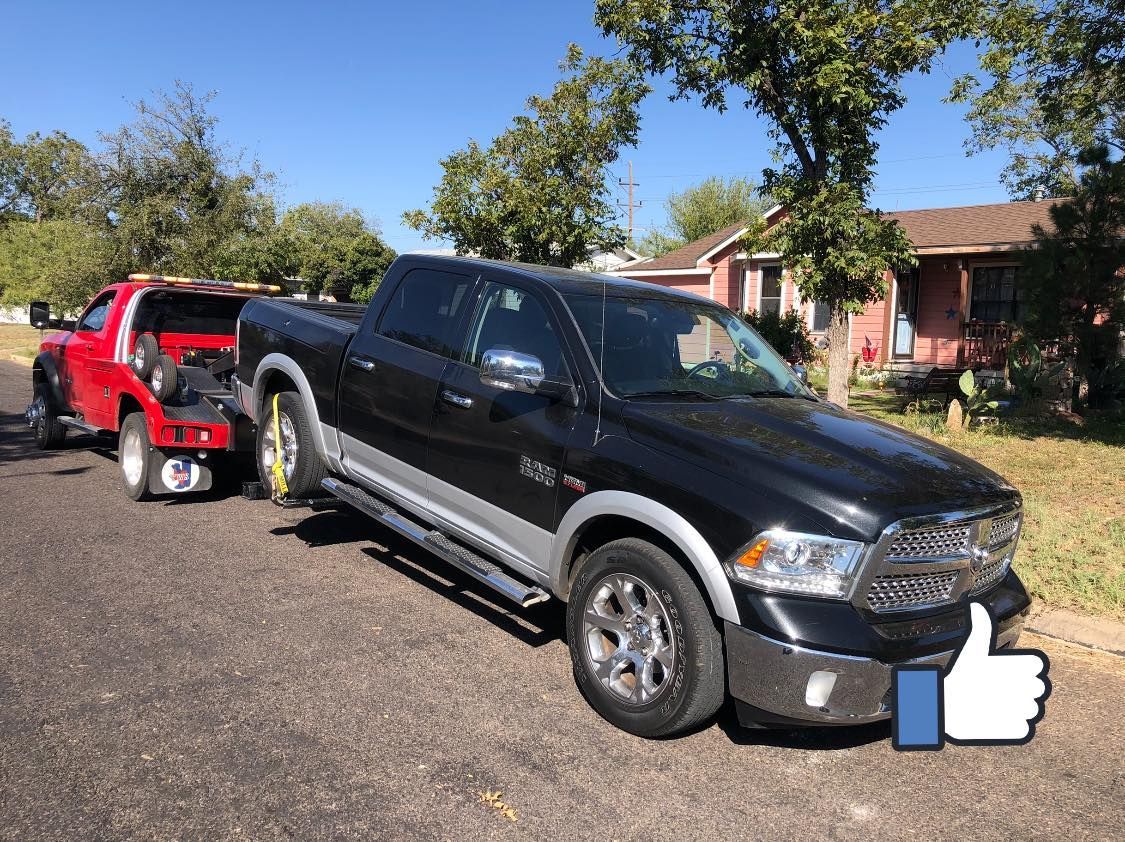 Black Ram truck being towed by a red tow truck on a street. Sunny day, residential setting.