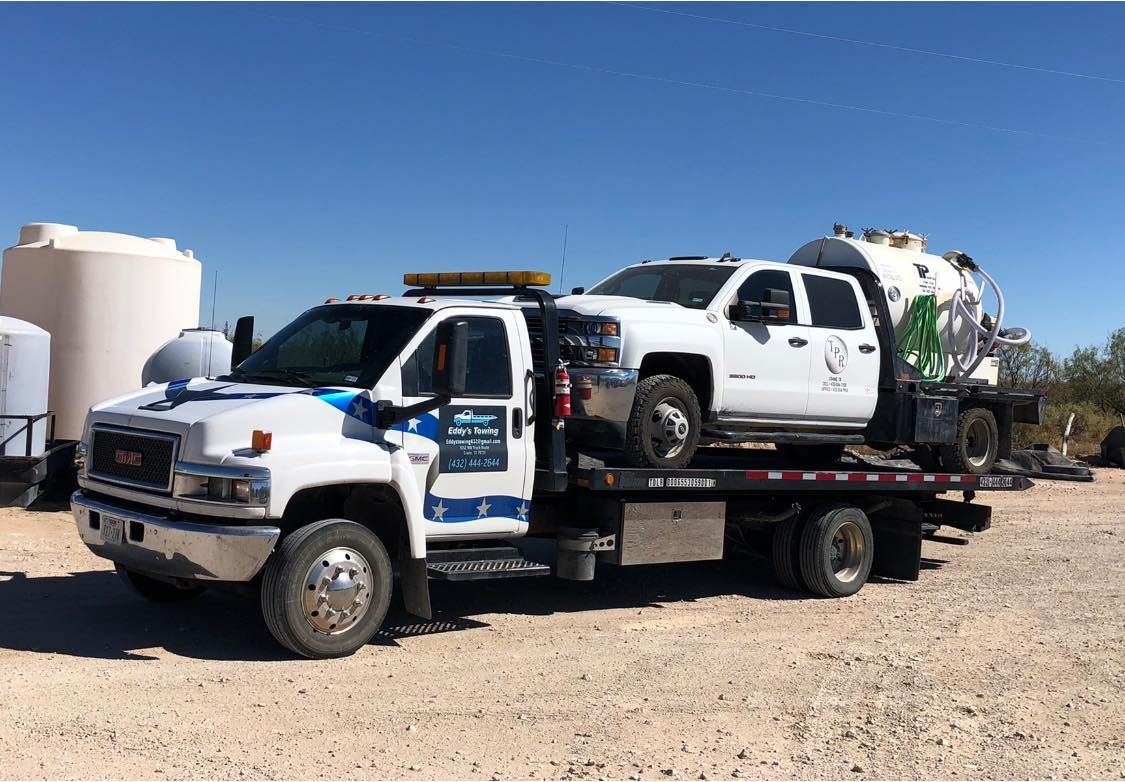 White tow truck carrying a white truck with a large tank, parked on dirt under a blue sky.