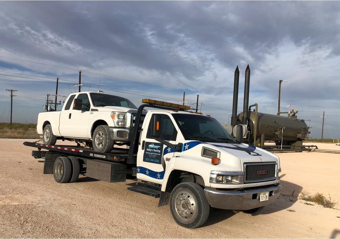 White tow truck hauling a white pickup truck on a gravel road, oil field in background.