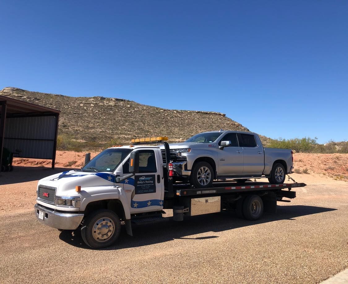 Tow truck with silver pickup truck on its flatbed, under a blue sky in a desert landscape.