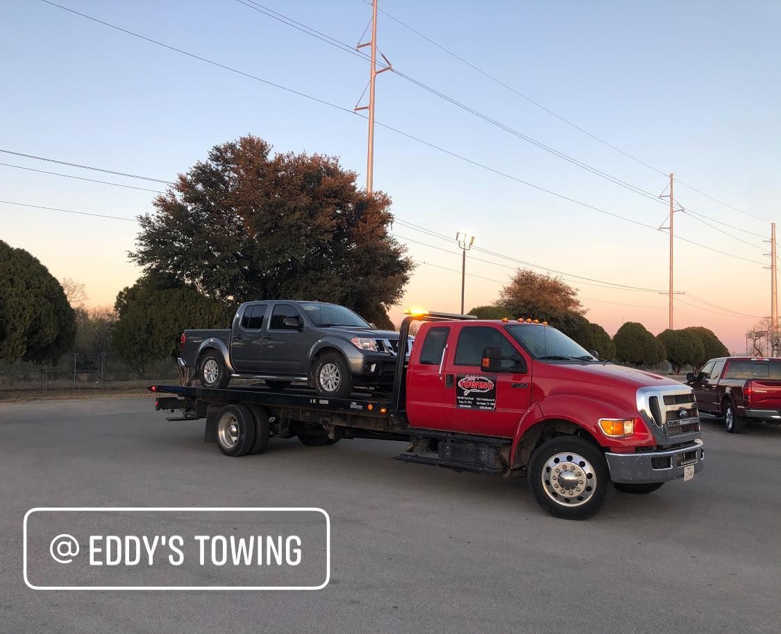 Red tow truck carrying a gray pickup truck on a road; sunset in background; 