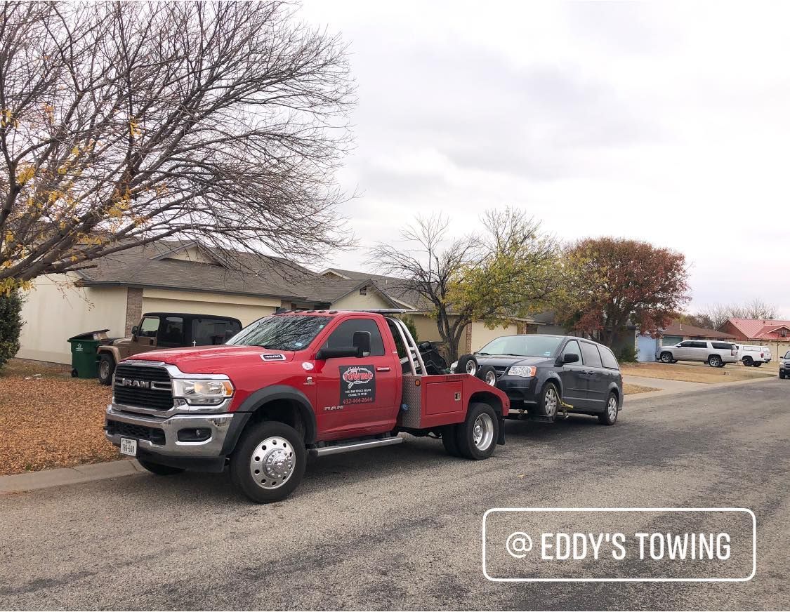 Red tow truck towing a gray SUV on a residential street. Cloudy sky, trees, and houses in the background.