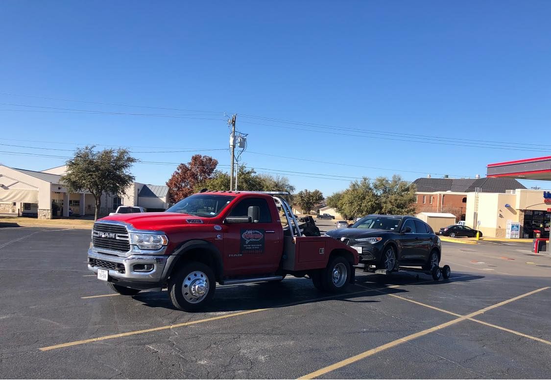 Red tow truck towing a dark SUV in a parking lot on a sunny day.
