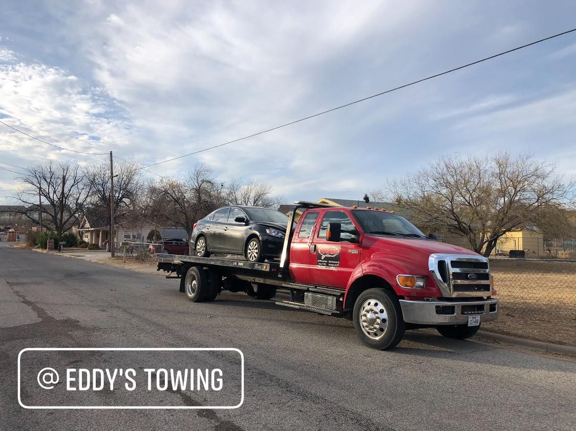 A red tow truck carrying a gray car on a street, cloudy sky overhead.