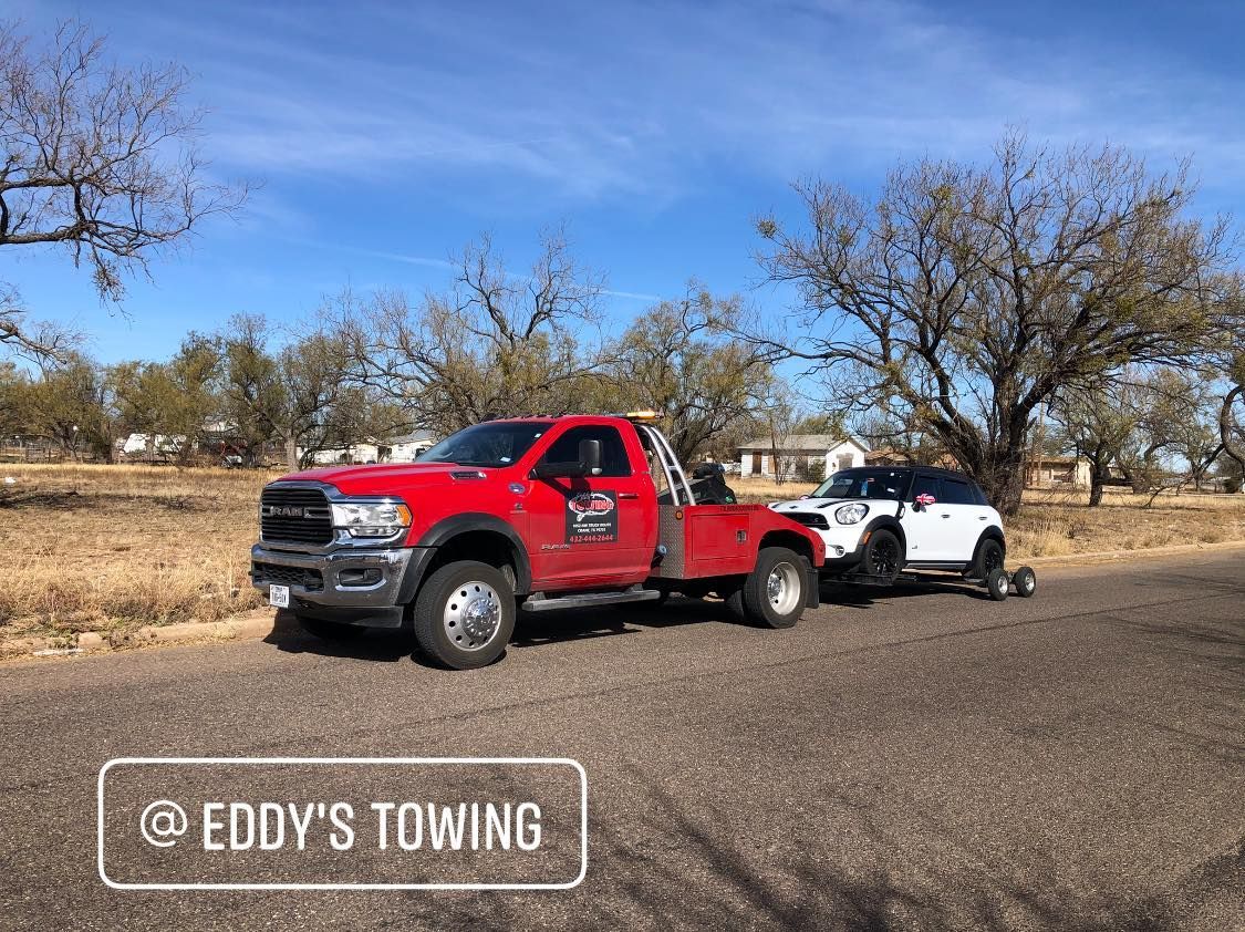Red tow truck towing a white SUV on a paved road under a blue sky, trees in the background.