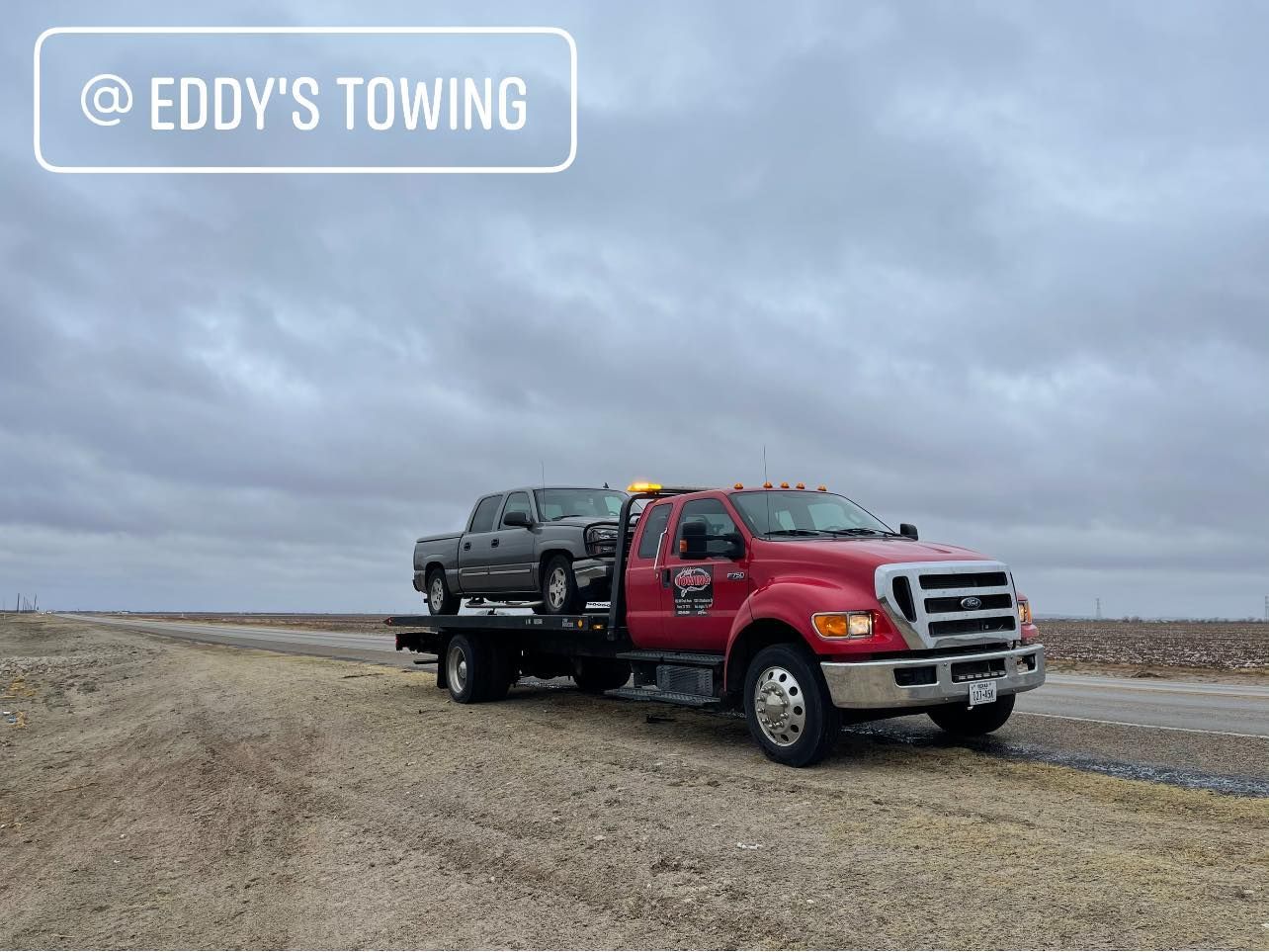 Red tow truck towing a gray pickup truck on a dirt road under a cloudy sky.