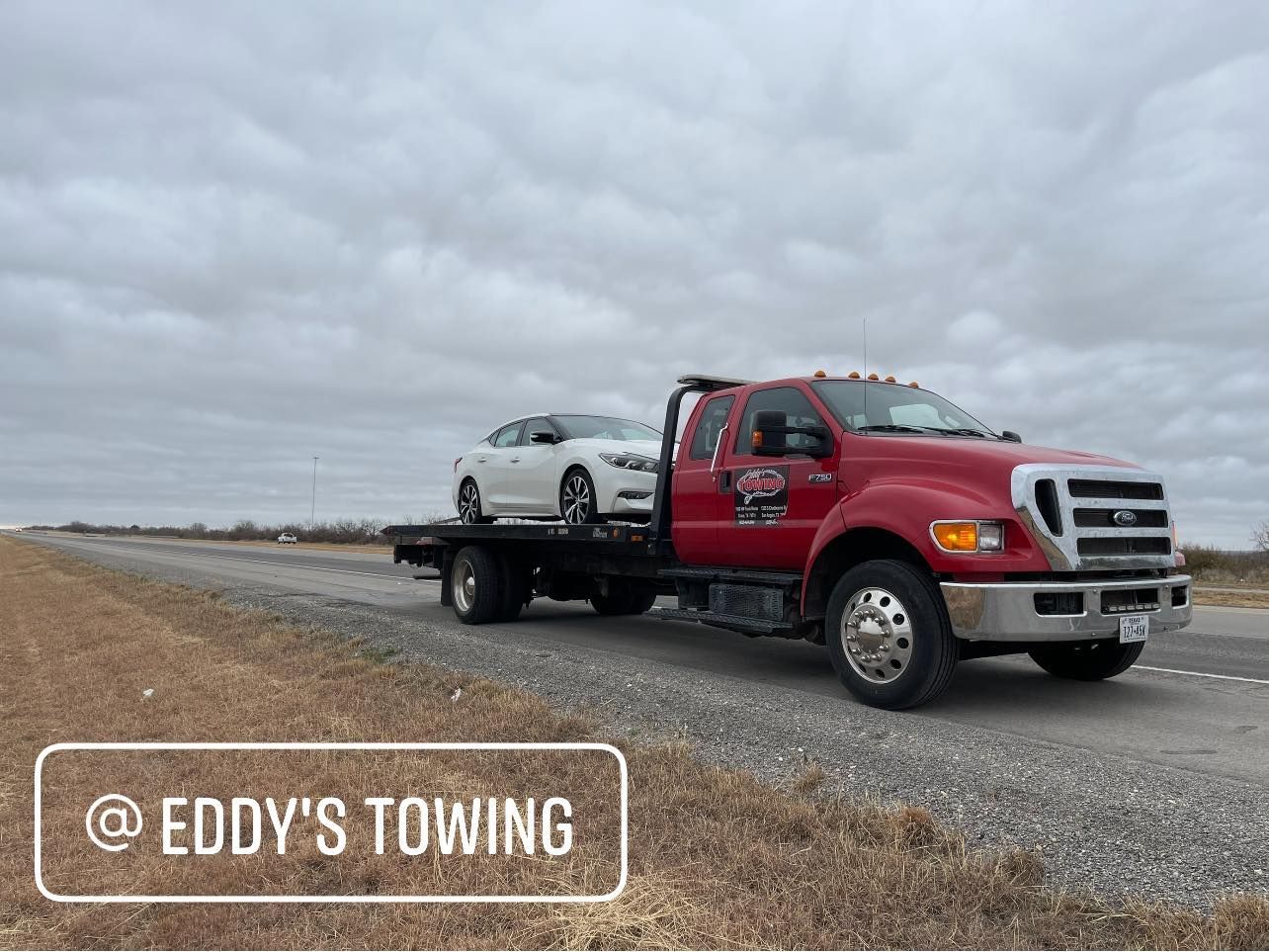 Red tow truck carrying a white car on a cloudy day. Roadside setting. 