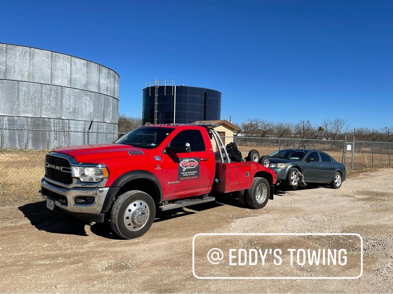 Red tow truck towing a gray sedan on a dirt road, near water tanks under a blue sky.