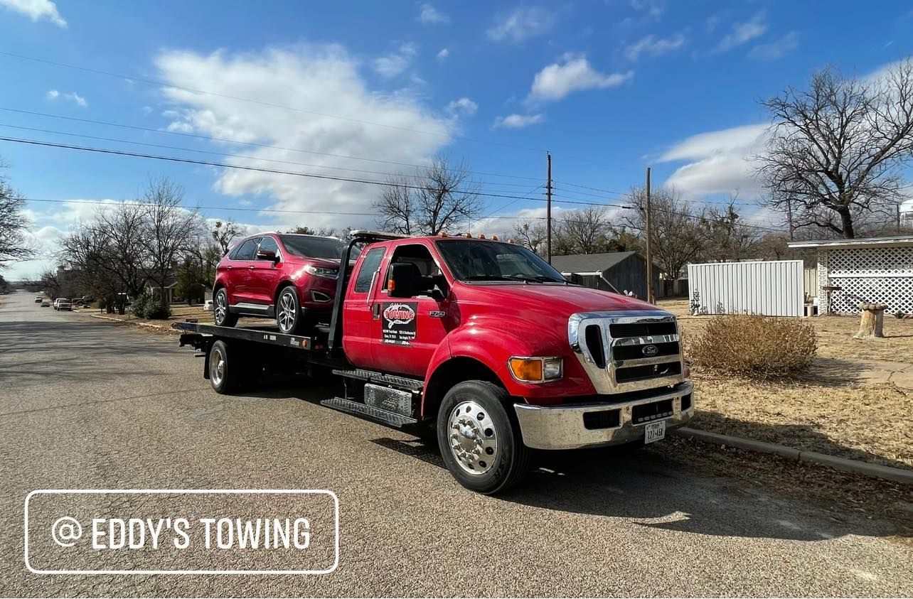 Red tow truck carrying a red car on a street; @ Eddy's Towing logo.