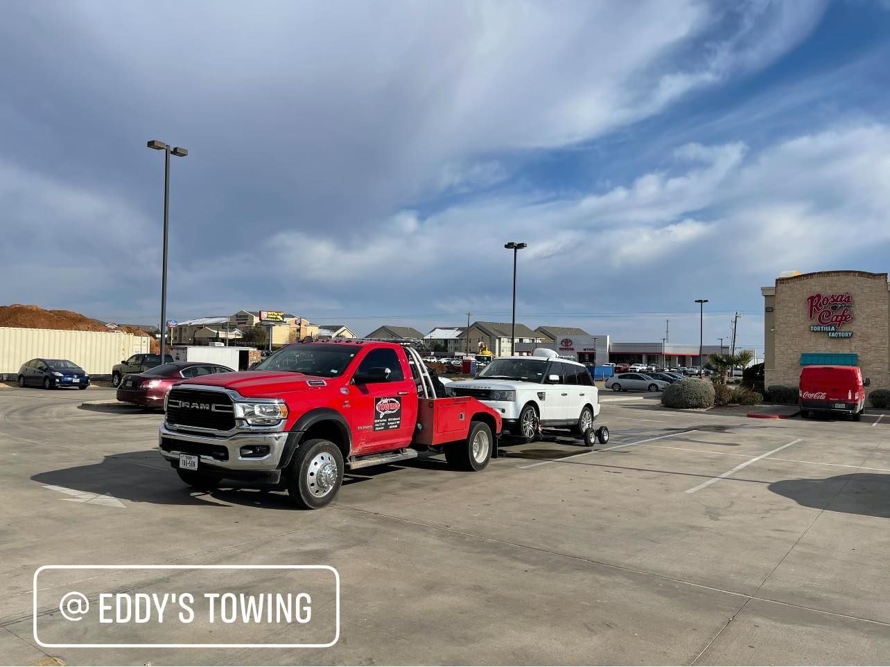 Red tow truck towing a white vehicle in a parking lot on a cloudy day.