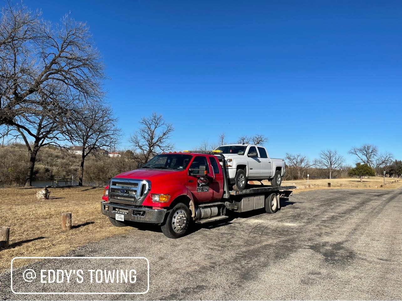 Red tow truck with a white pickup on its flatbed, parked outdoors under a blue sky.