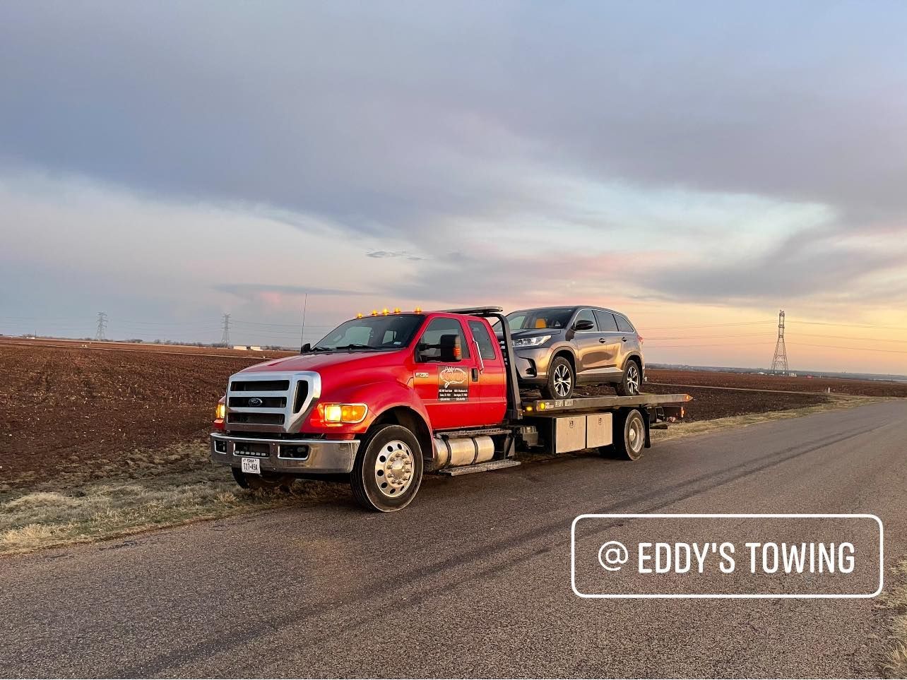 Red tow truck with a silver SUV on a flatbed, on a rural road at sunset; 
