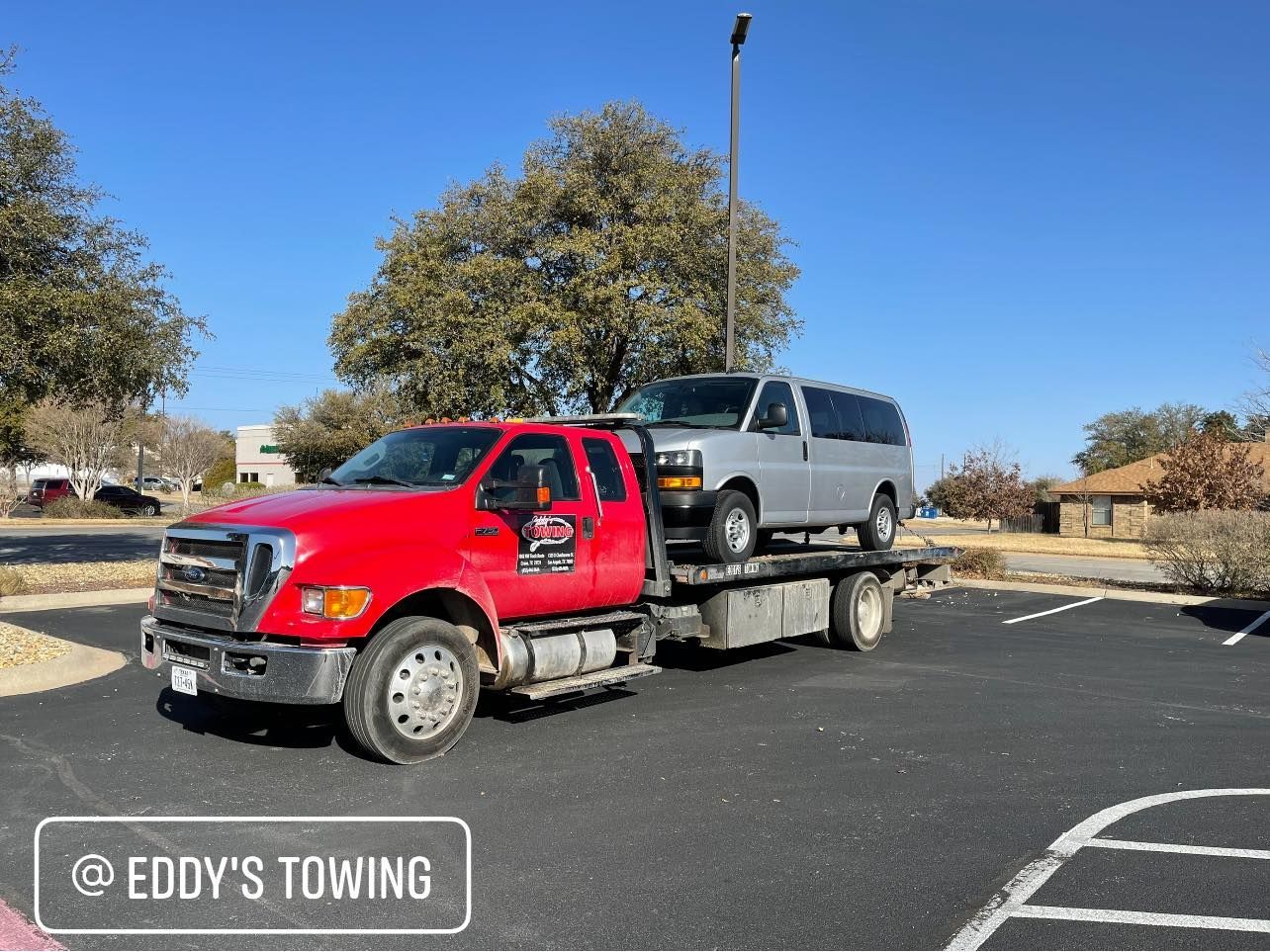 Red tow truck hauling a silver van in a parking lot on a sunny day.