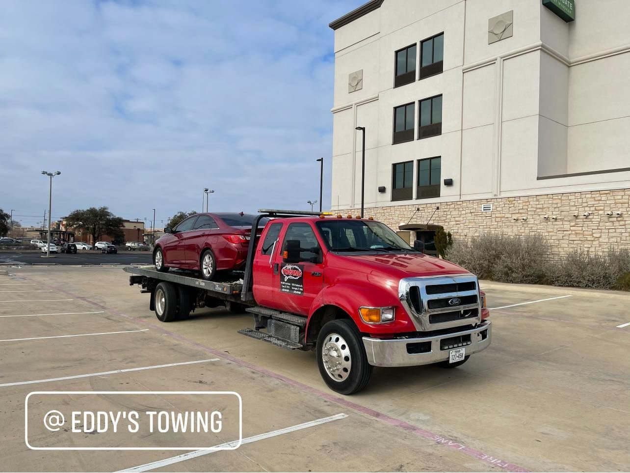 Red tow truck with a car on the flatbed in a parking lot. Building in the background.