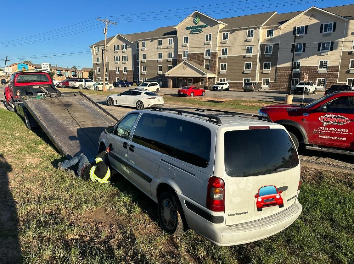 A tow truck is loading a white minivan from a grassy area near an Express Hotel on a sunny day.
