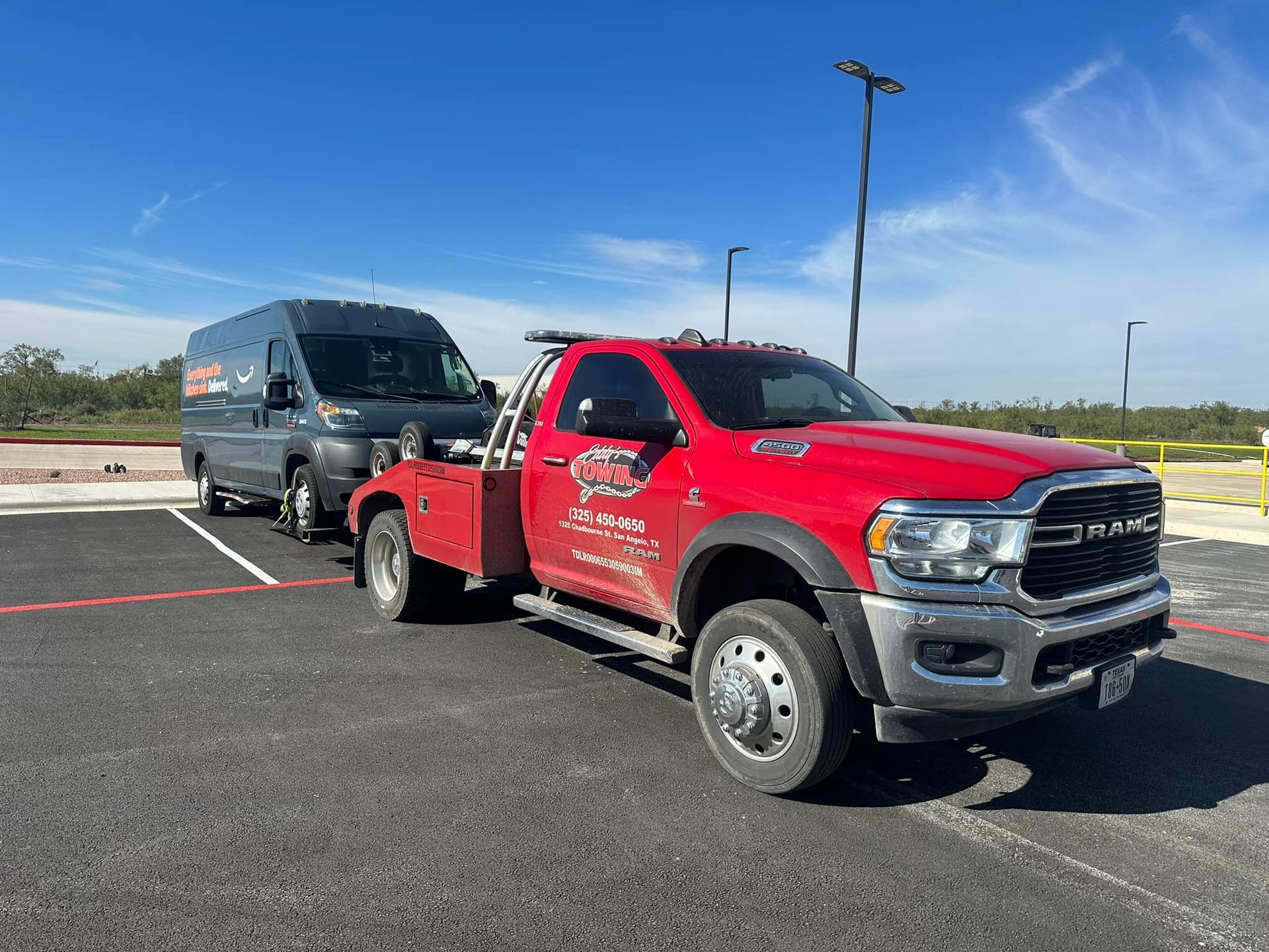 Red tow truck towing a gray van on a paved lot under a clear sky.